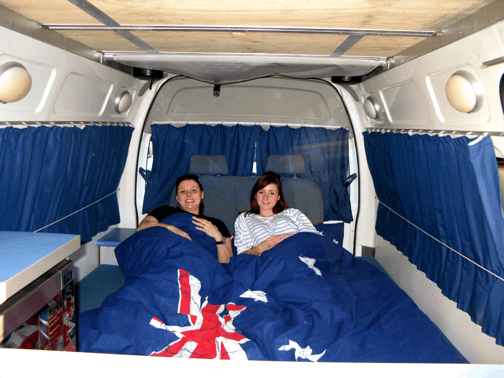 Photo of big double bed with two girls under the blankets inside this Automatic campervan hire