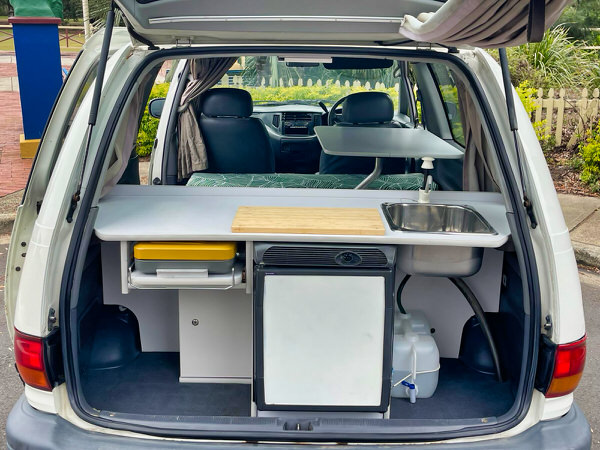 Toyota Tarago campervans for sale in Sydney - Automatic 2 person campervan model. Photo shows the view of the rear tailgate open and the mini kitchen with fridge, cooker, sink, water tank and worktop space